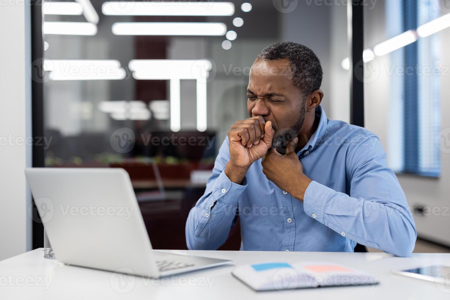 Mature African American businessman sitting at desk in modern office, coughing into hand. Concerned expression indicates health issues affecting work. photo