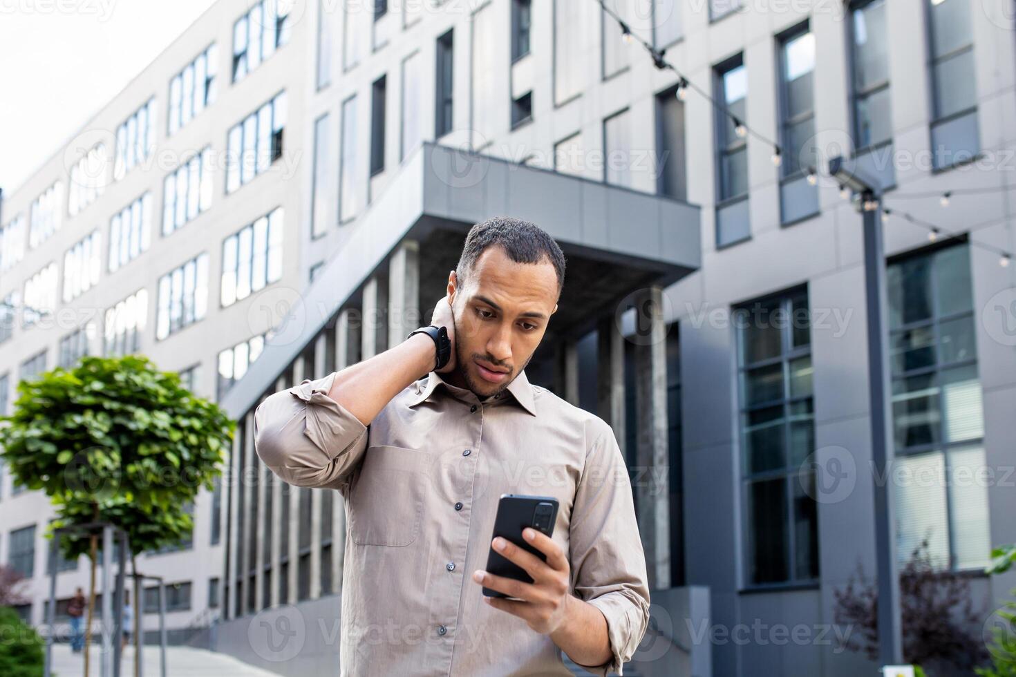 Disturbed arab man rubbing head with hand while looking at mobile phone screen on background of office building. Stressed manager leaving corporate workplace and receiving text message from boss. photo