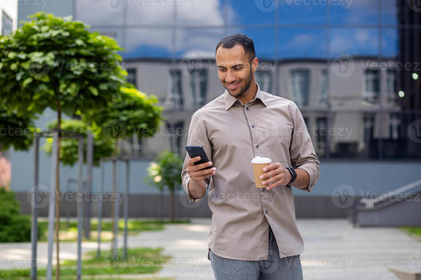 Relaxed middle eastern male having walk by corporate building exterior with plastic coffee cup and cellphone. Happy formal manager enjoying break with hot beverage from work and warm weather outdoors. photo