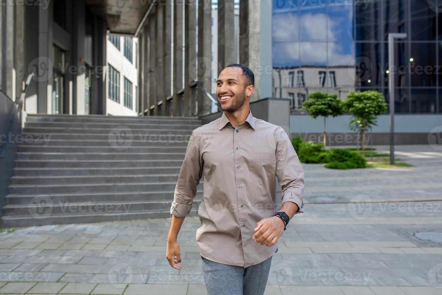 Energetic muslim male agent walking down street with satisfied facial expression. Optimistic agent leaving office building after profitable productive day and feeling excited about weekend ahead. photo