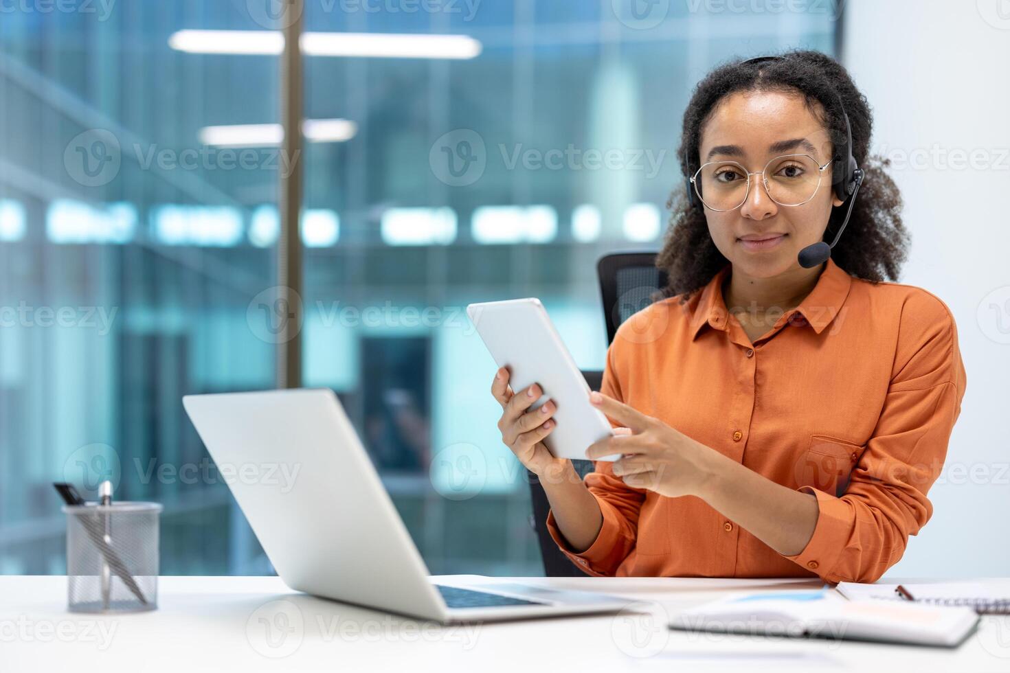 A young professional woman works in a modern office using a tablet and headset, while a laptop sits on her desk photo