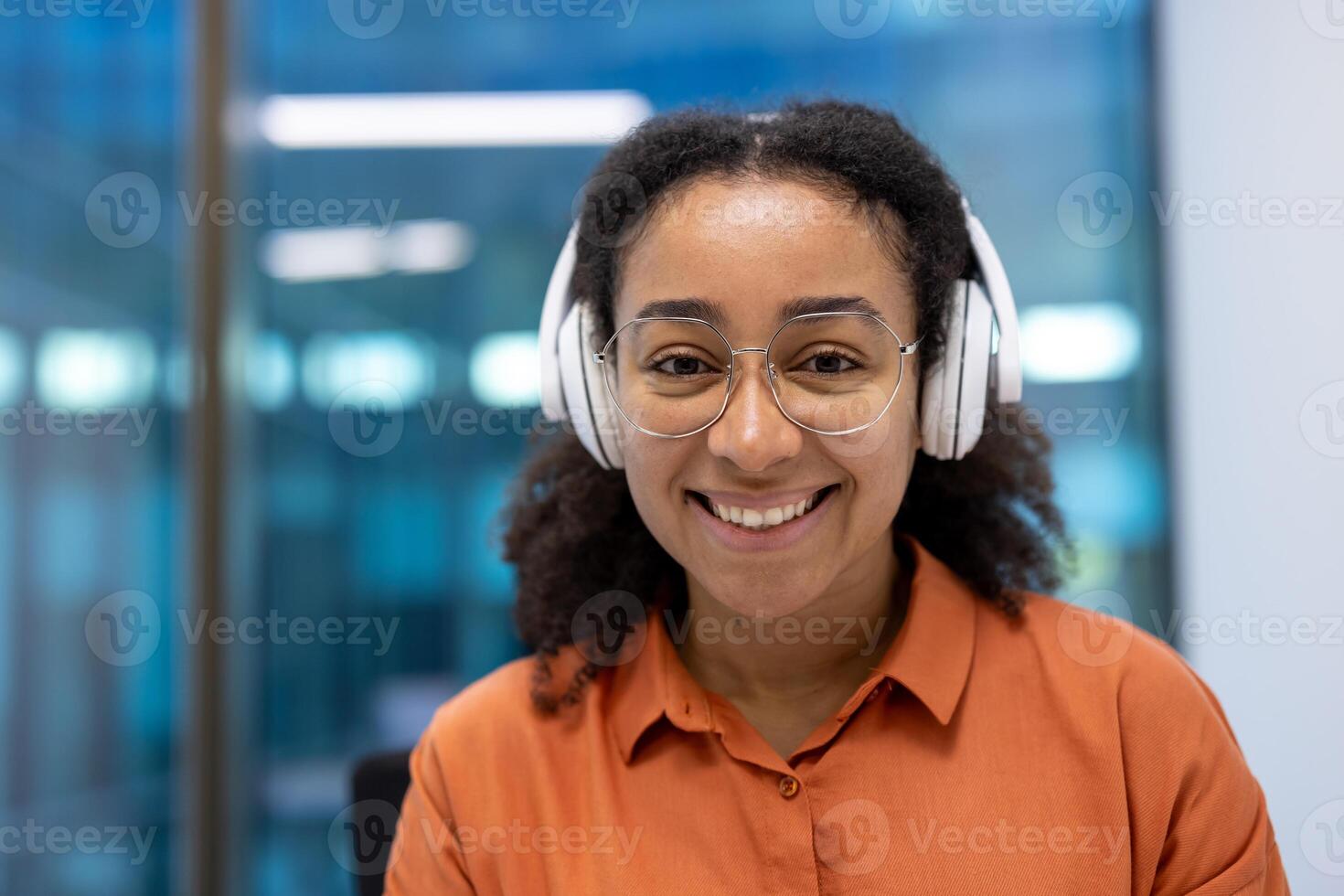 An attractive woman wearing headphones and glasses looks directly at the camera with a warm smile. She wears an orange top. photo