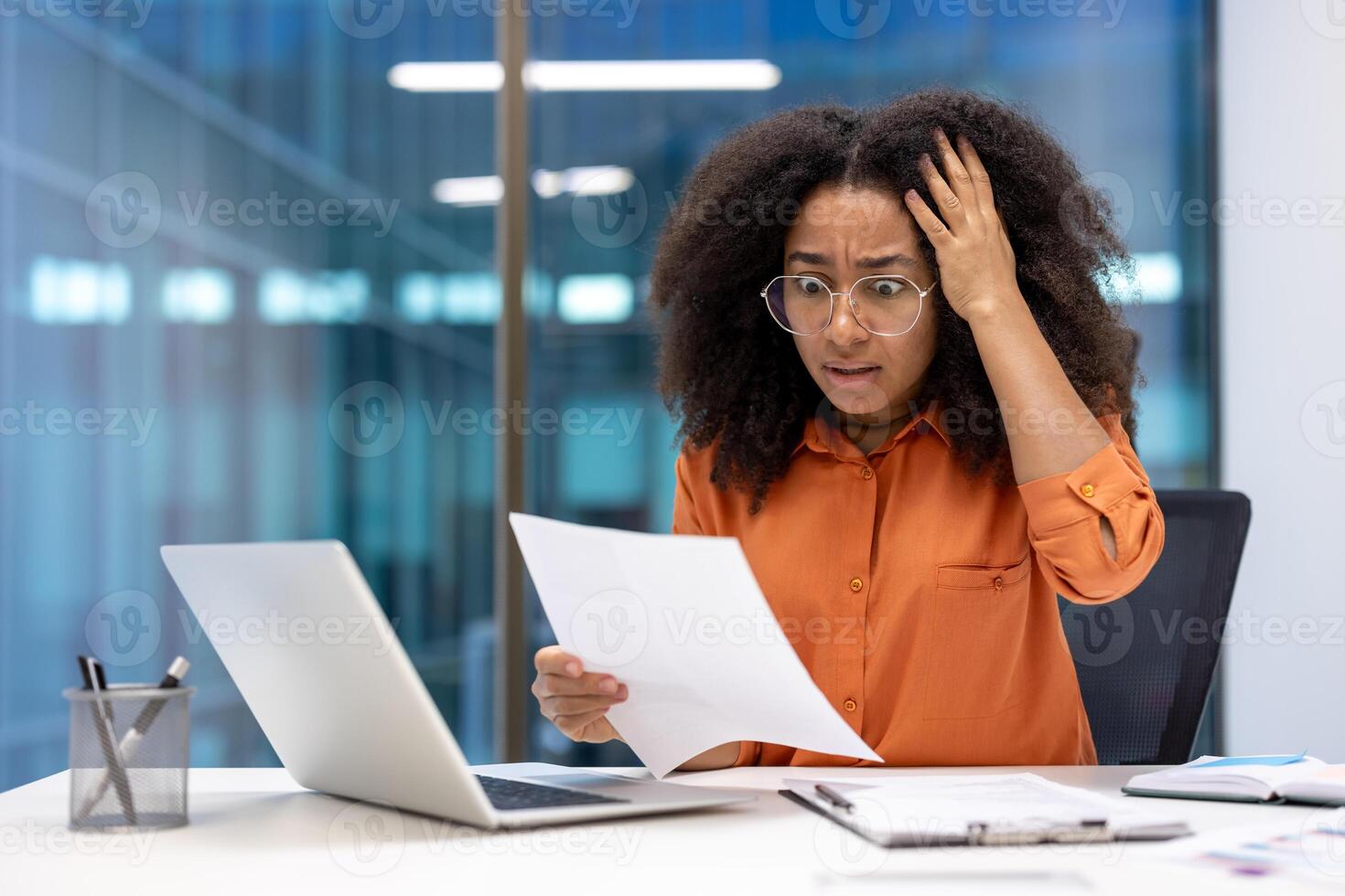 A businesswoman expresses worry while looking at a document. She is seated at a desk with a laptop, papers, and a pen holder in front of her. photo