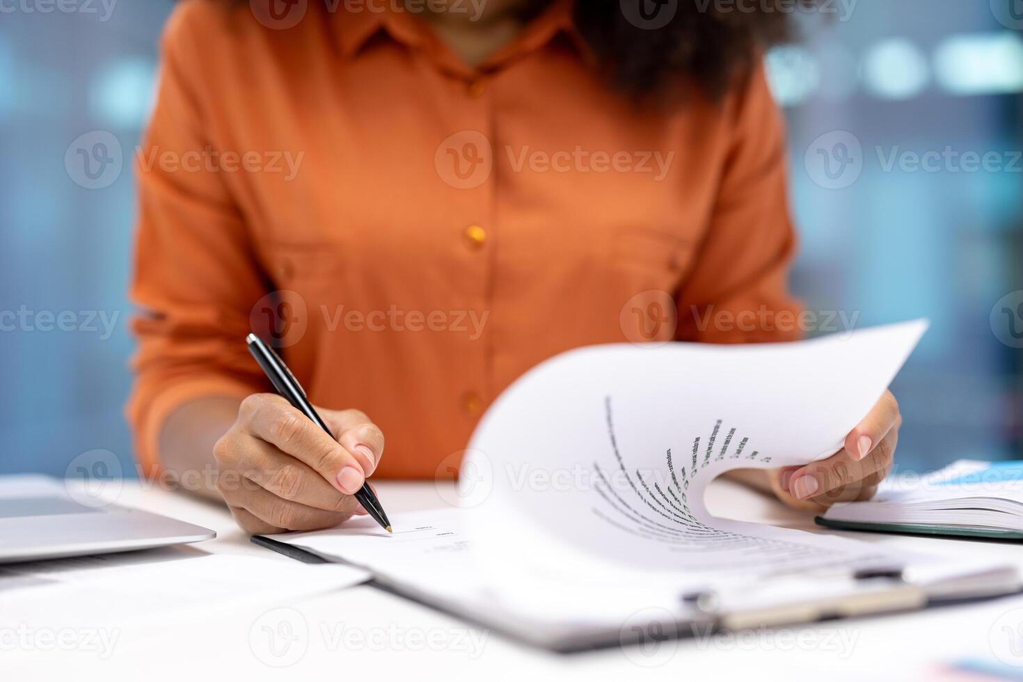 A person in an orange shirt is writing on a document in an office setting, focusing on work with pen and paper. photo