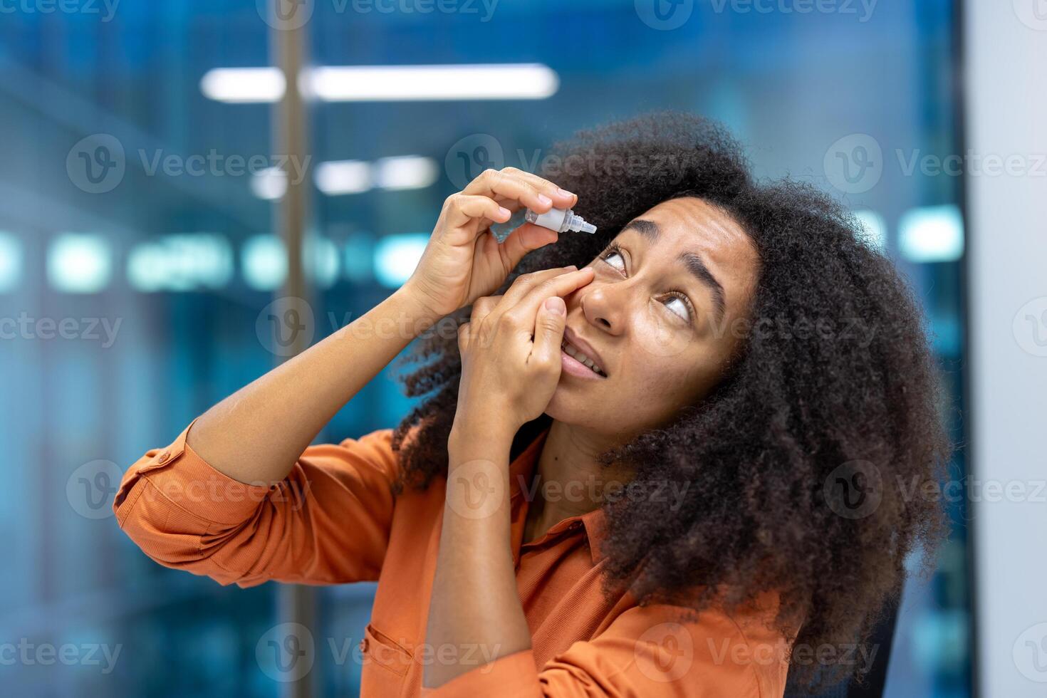 A woman with curly hair uses eye drops, likely to relieve irritation or dryness, indoors with natural light. photo