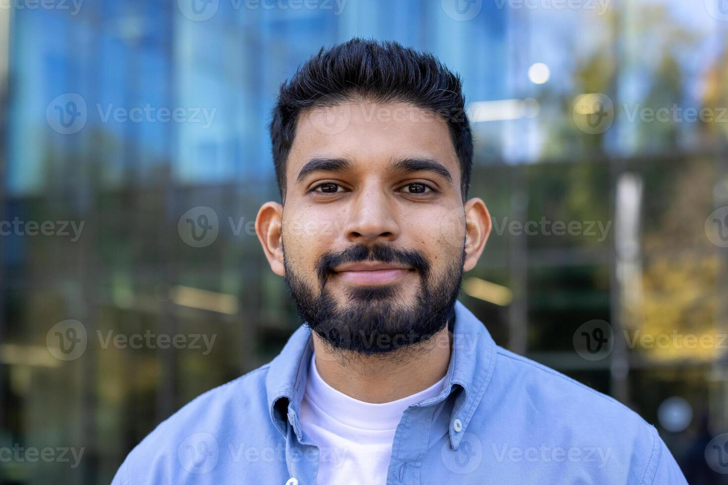 A handsome man with a beard smiles directly at the camera while standing outside. The background shows a modern building with glass windows. photo