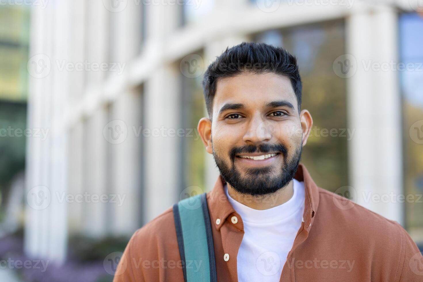 A smiling young Indian man with a backpack stands outdoors, looking at the camera. He is wearing a shirt and smiling. photo