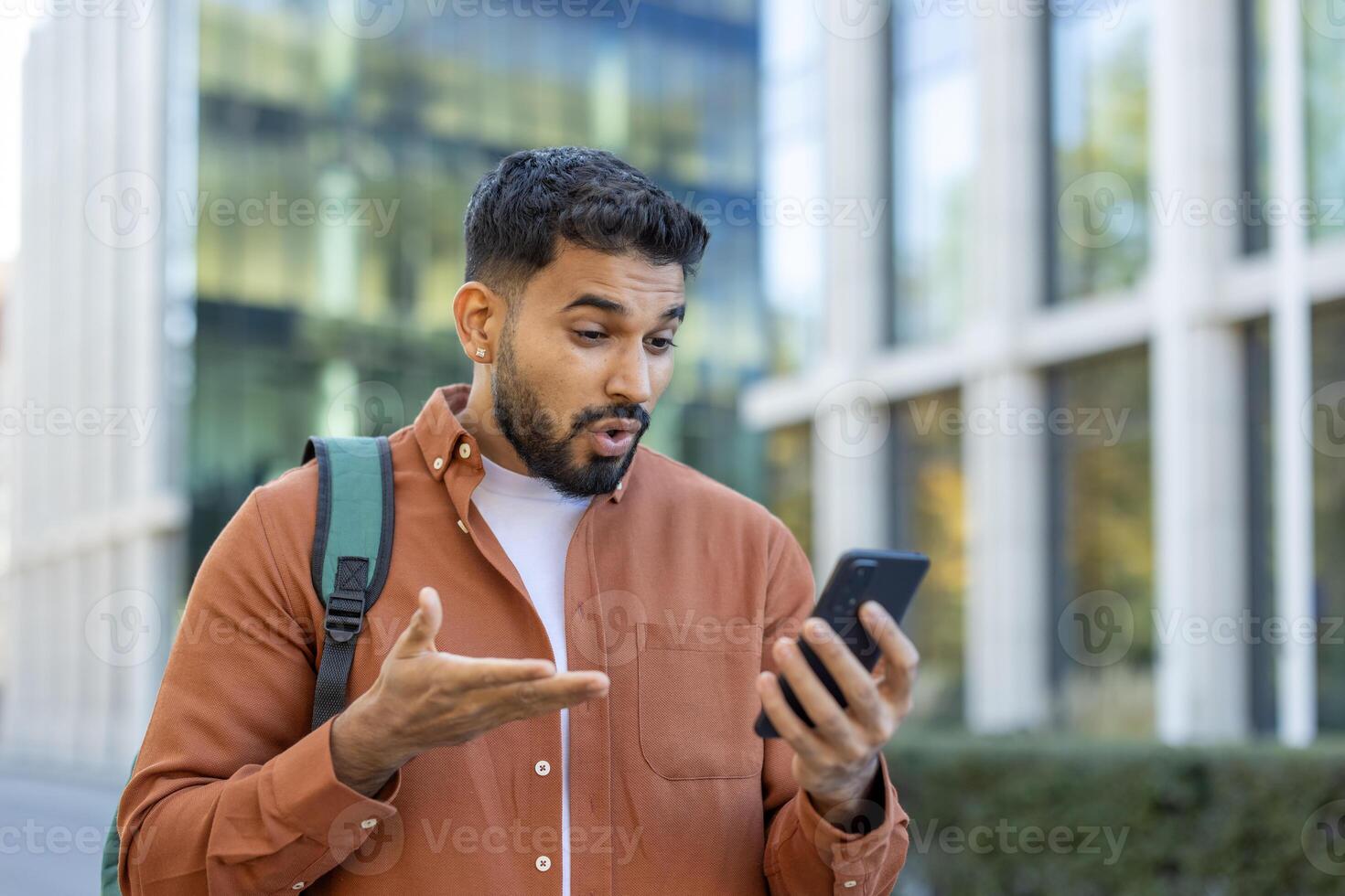A young man outside reacts with surprise while looking at his smartphone. He gestures with his hand, seemingly in shock at the news displayed on the screen. photo