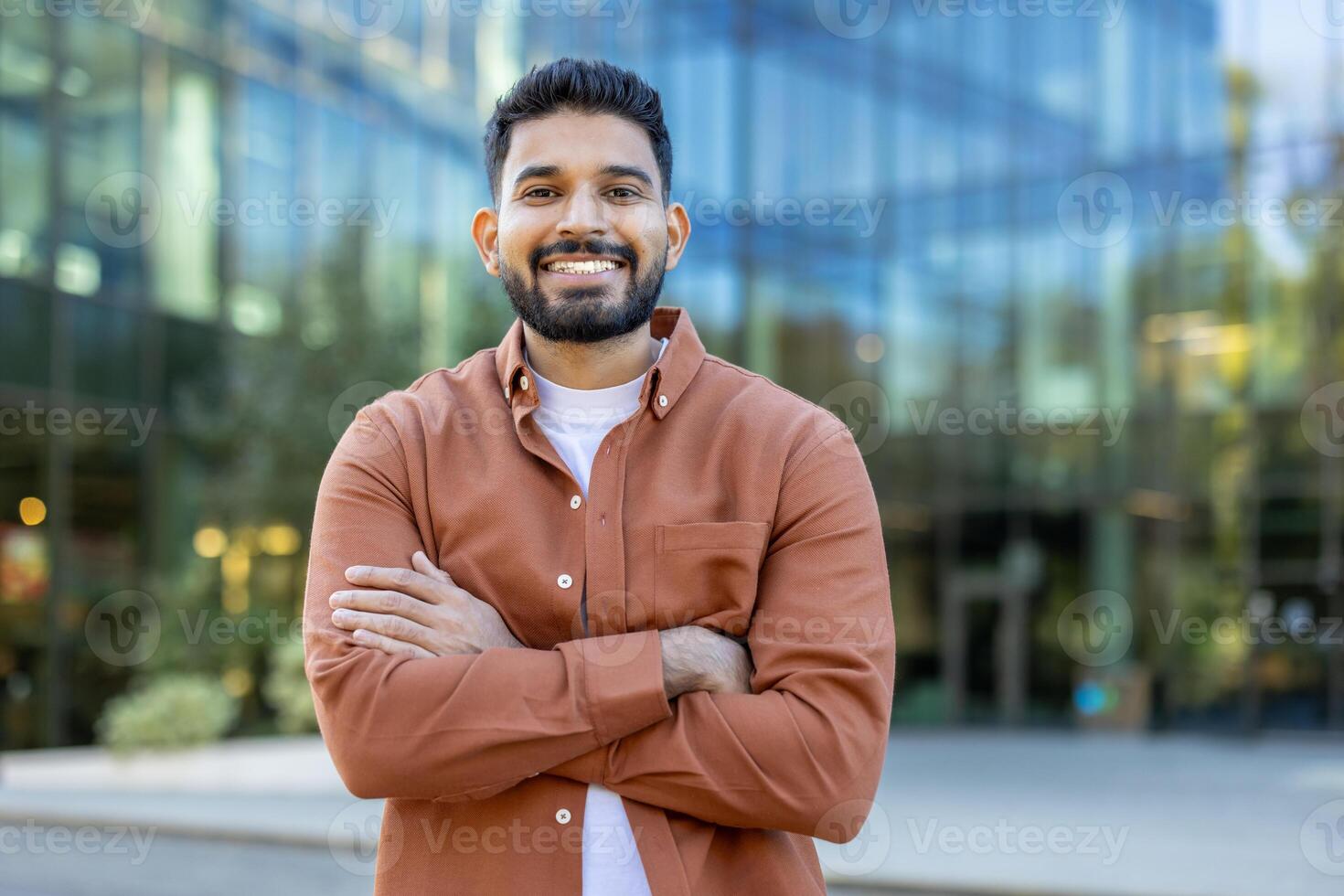 A smiling man stands confidently with arms crossed, posing in front of a modern glass building. The outdoor setting captures a professional and approachable vibe. photo