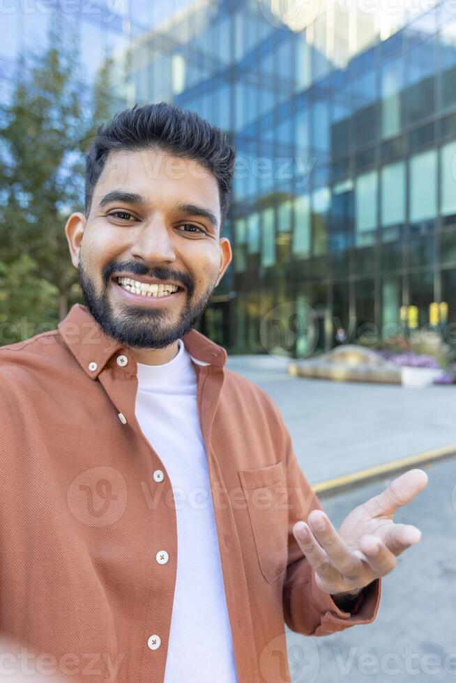 A man smiling and gesturing with his hand during a call with a modern building in the background. He is friendly photo