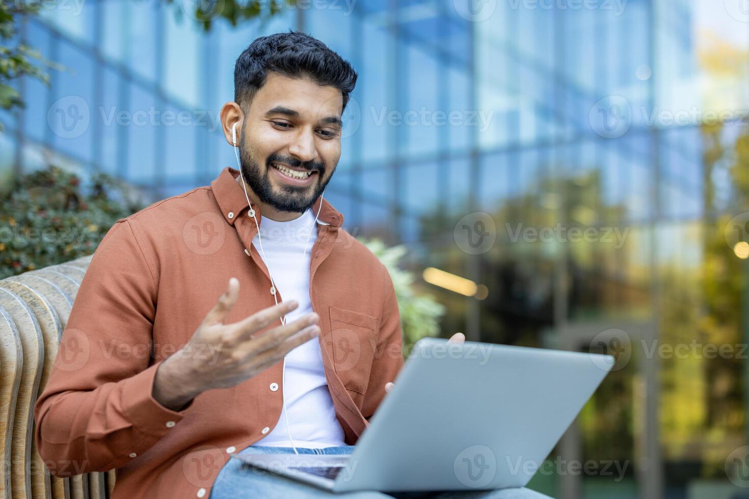 A smiling man in casual wear attends a call, using a laptop and wired earbuds while seated outdoors, with a modern building backdrop. photo