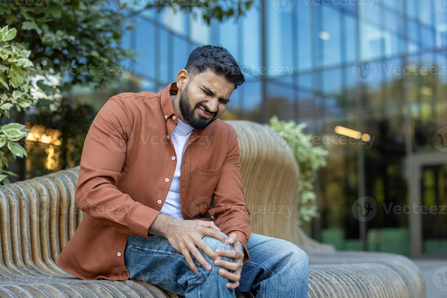 A man is sitting on a bench, visibly experiencing knee pain, his expression showing discomfort and strain. The photo is taken outdoors, during the day.
