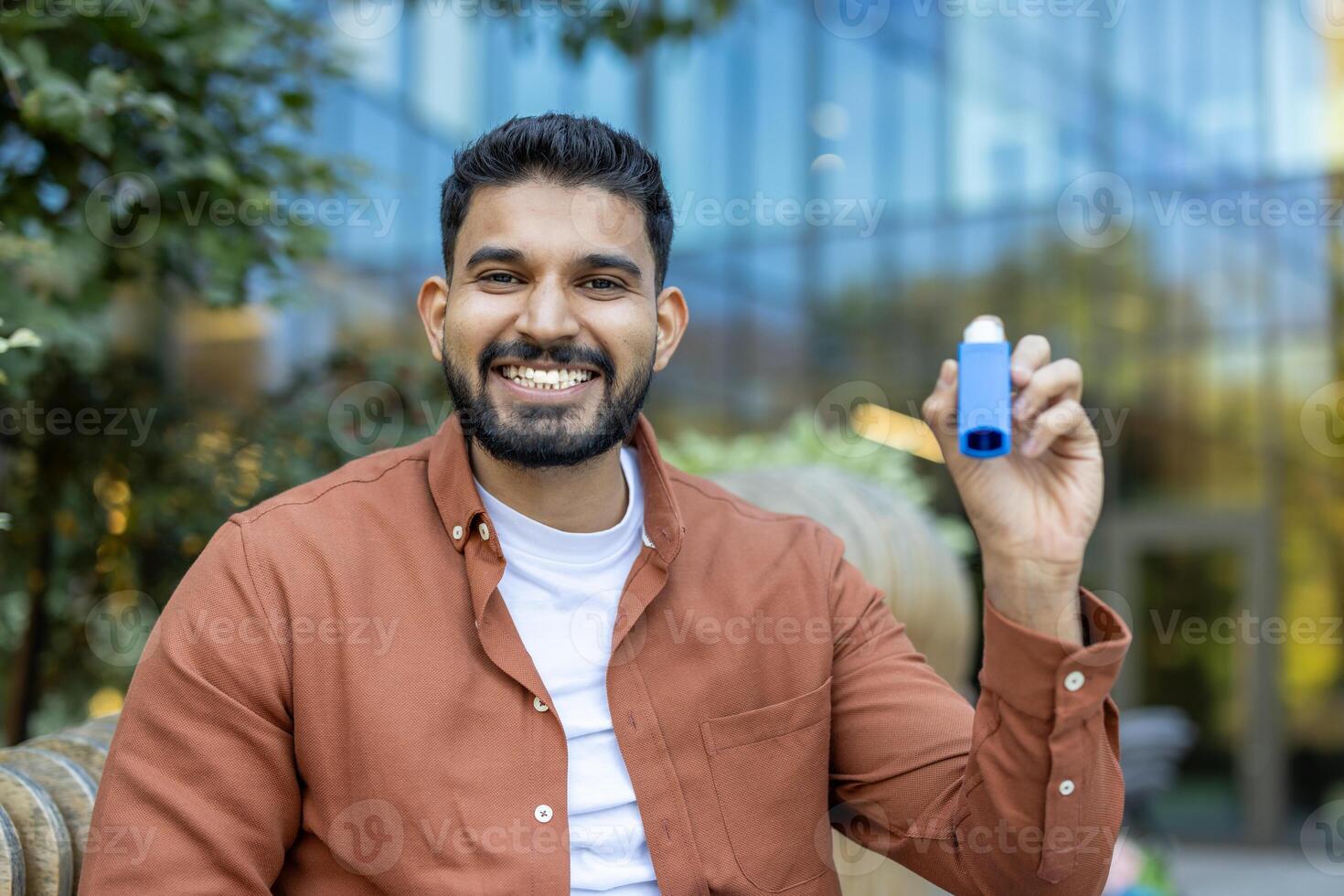 A smiling man outdoors holds up an inhaler, indicating asthma or respiratory health. The background shows a blurred building and greenery. photo