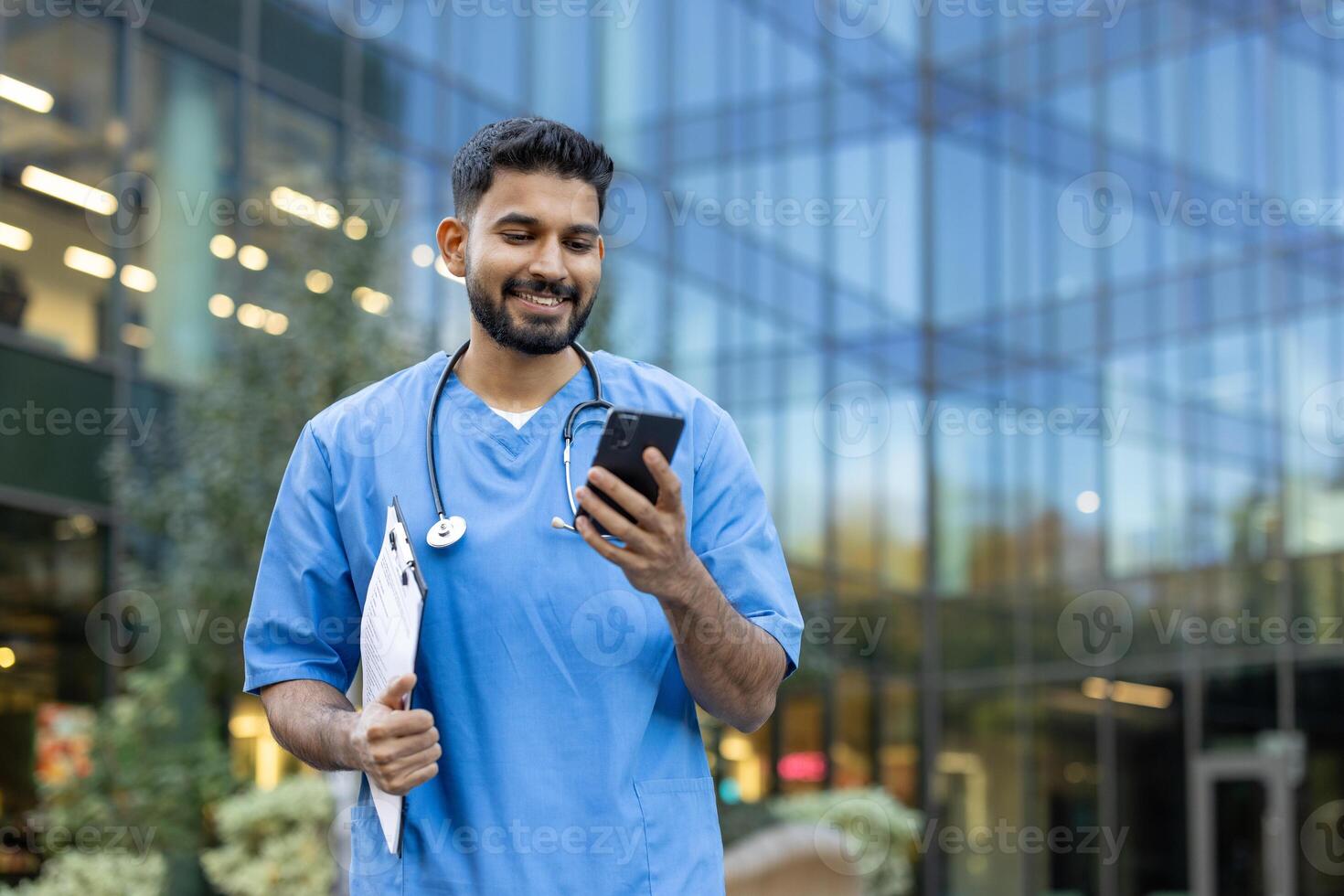 A medical professional looks at his smartphone, smiling, while outside near a modern building. He's holding a clipboard and a stethoscope. photo