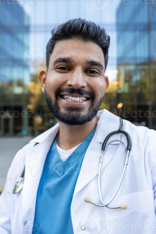A friendly doctor stands outdoors, wearing a white coat and stethoscope, smiling brightly at the camera. The background includes a modern building. photo