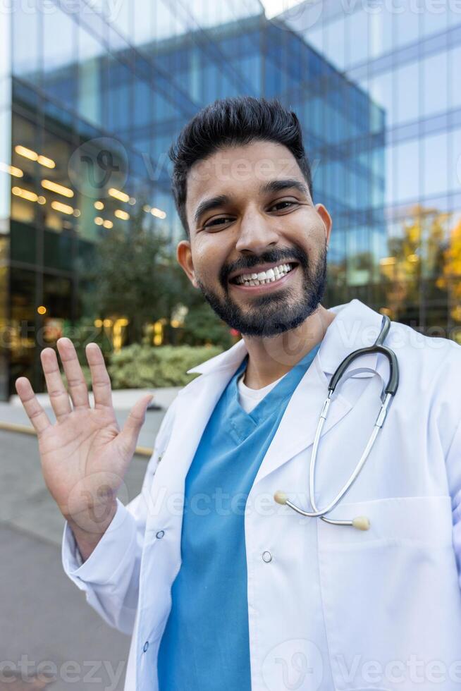 A smiling doctor waves at the camera outside a modern building, wearing a stethoscope. He is welcoming and approachable in his medical attire. photo