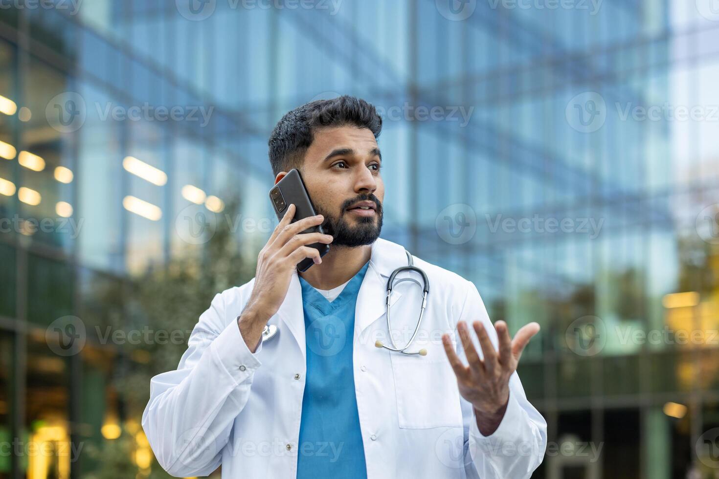 A doctor wearing a white coat speaks on his phone outdoors, likely consulting with a colleague or patient, near a modern building. photo