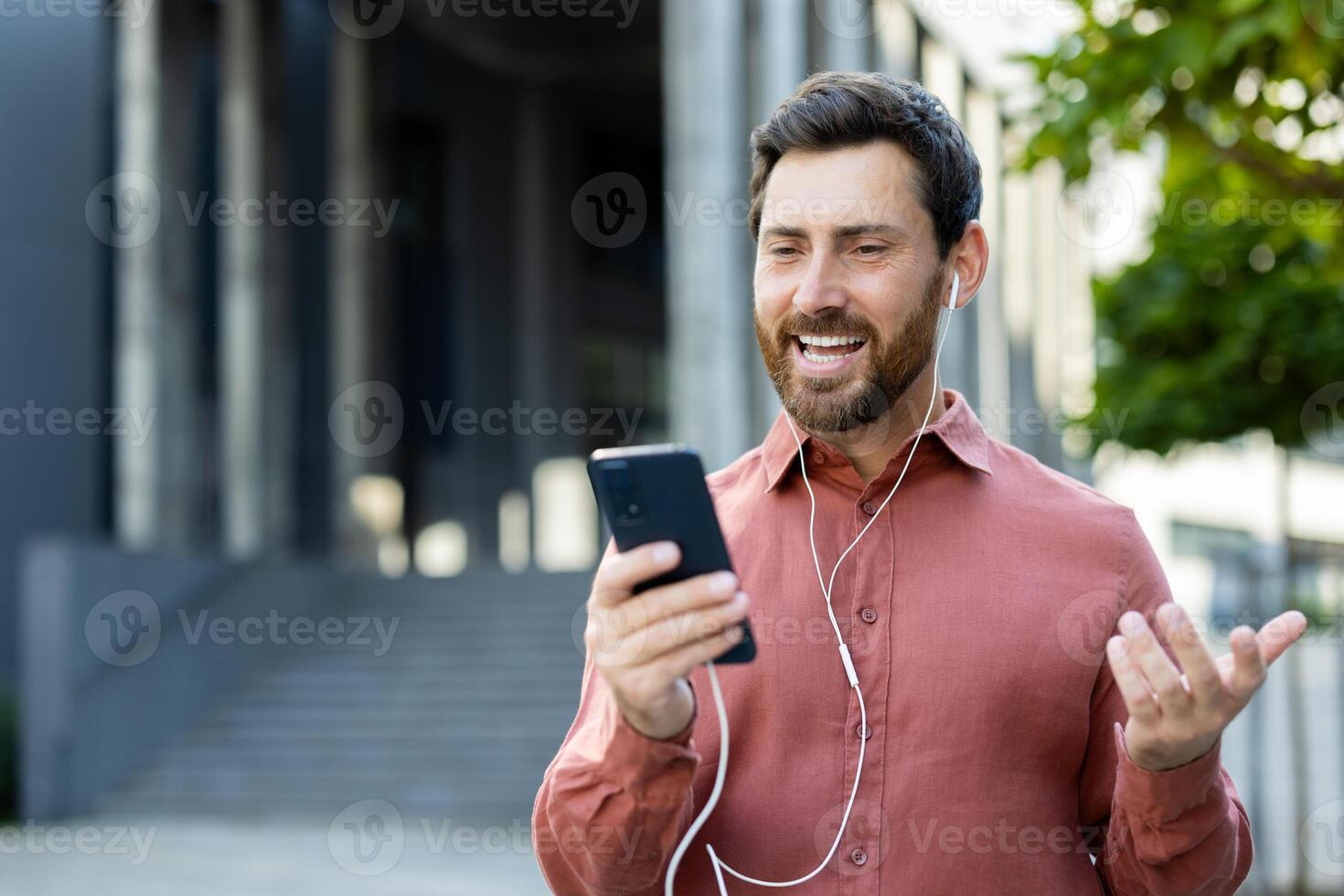 A man in a red shirt is having a call while outside of a building. He is smiling and gesturing. photo