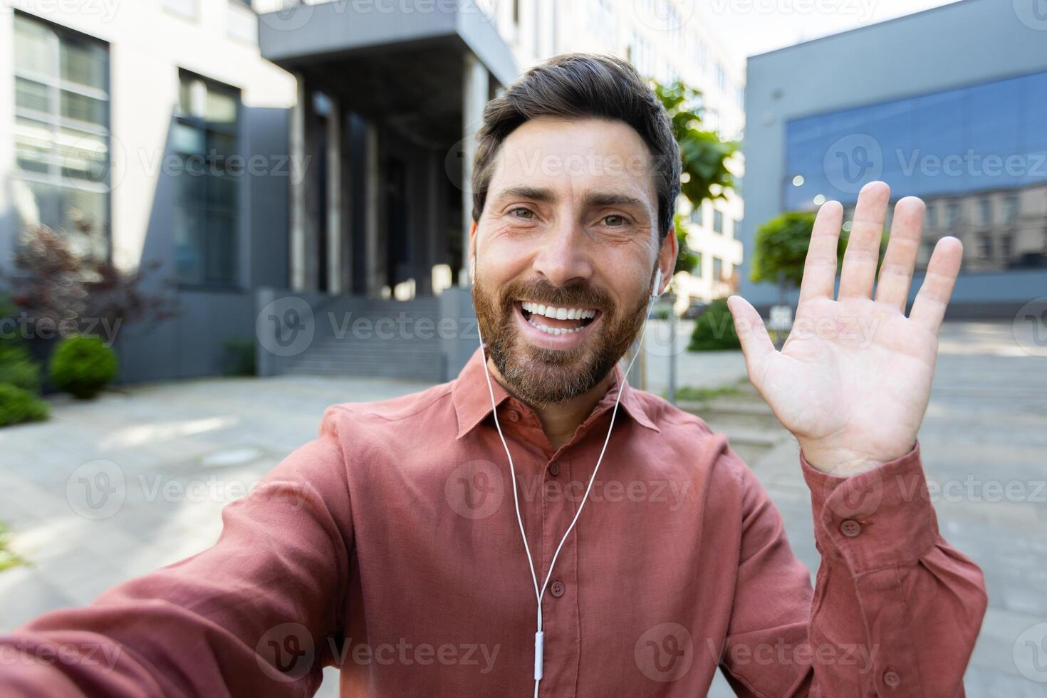 A man with a beard smiles while waving at the camera outside near a modern building, he wears earbuds. photo