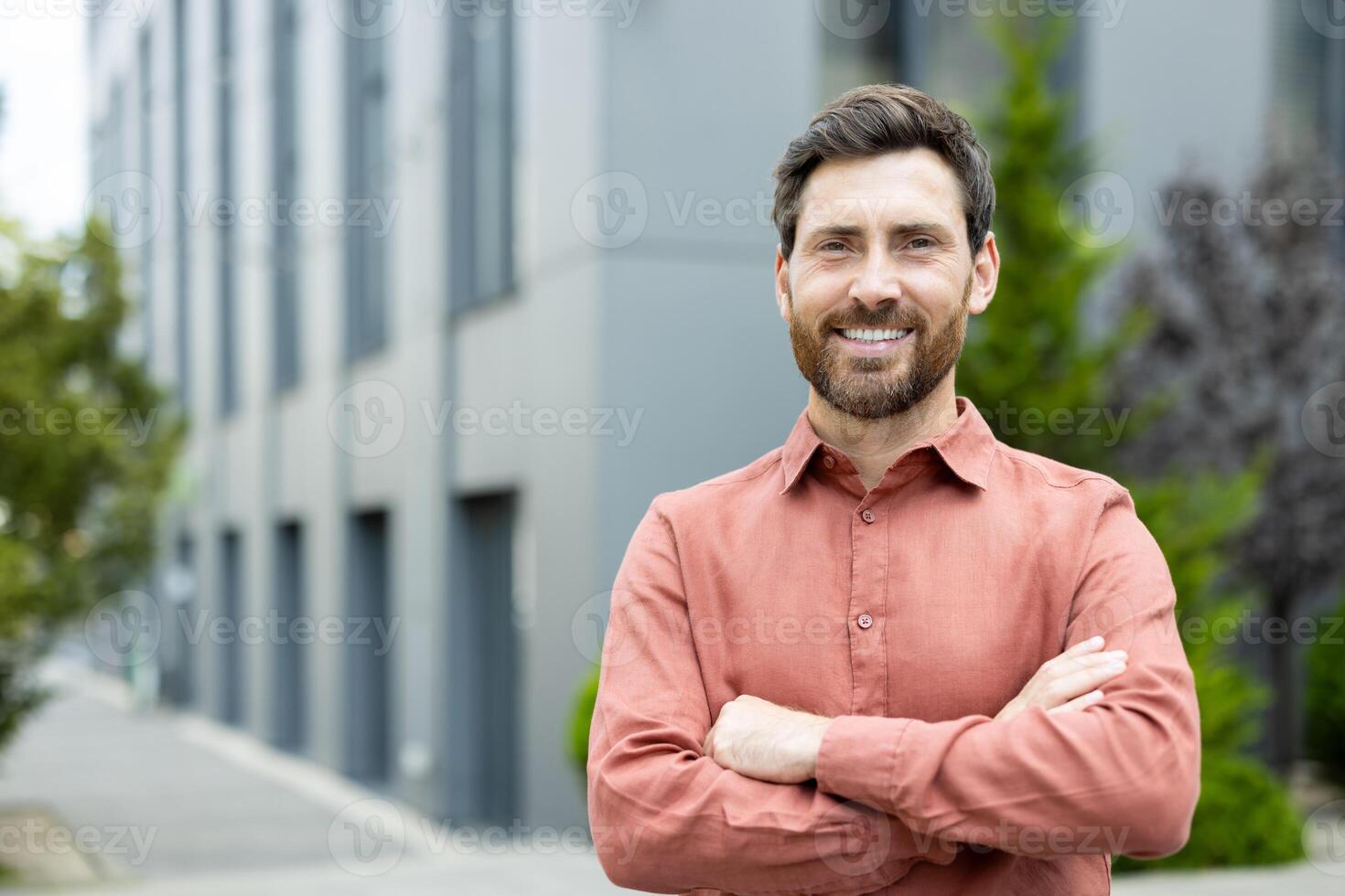 A smiling man in an orange shirt stands in front of a modern building, looking confident with arms crossed. photo