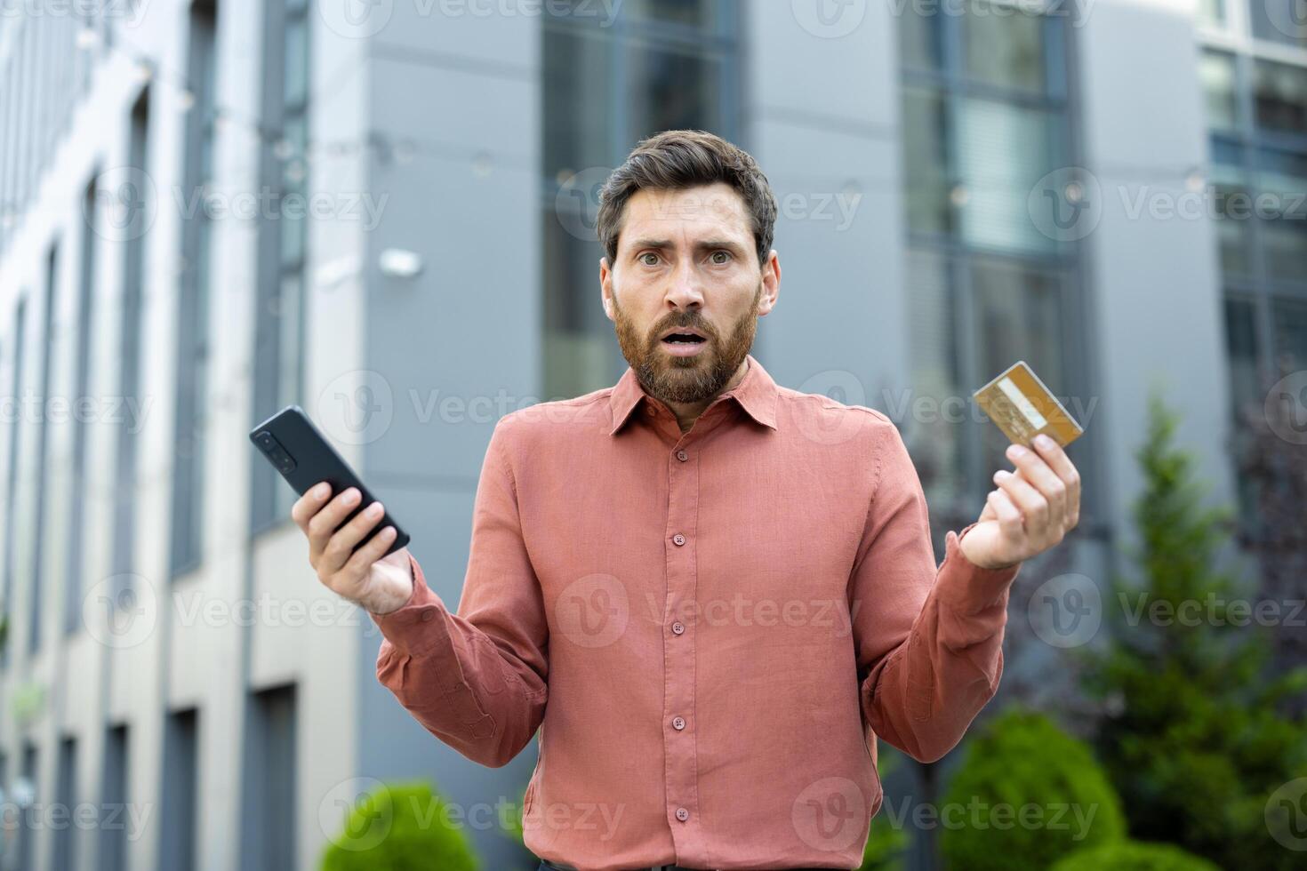 A man with a worried expression holds a credit card and a smartphone, seemingly facing a financial issue outdoors. photo
