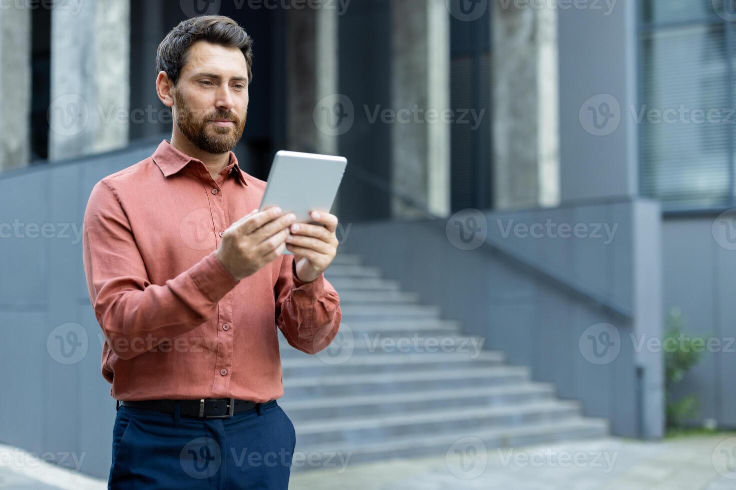A professional man in a button-up shirt looks at a tablet in front of a modern building. He could be checking emails or reading news. photo