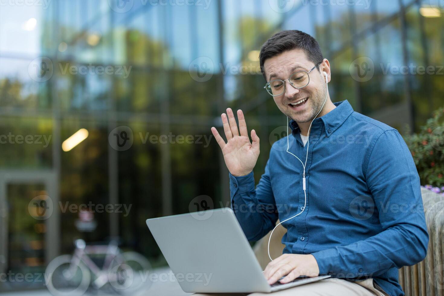 A man is sitting outside, using a laptop while waving to someone. He is smiling and appears to be having a call. photo