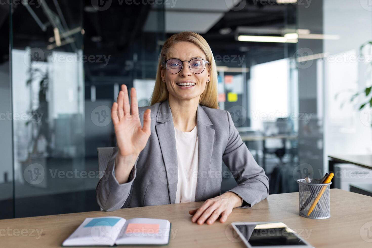 A businesswoman waves during a conference, wearing glasses and a grey suit jacket, with desk items visible photo