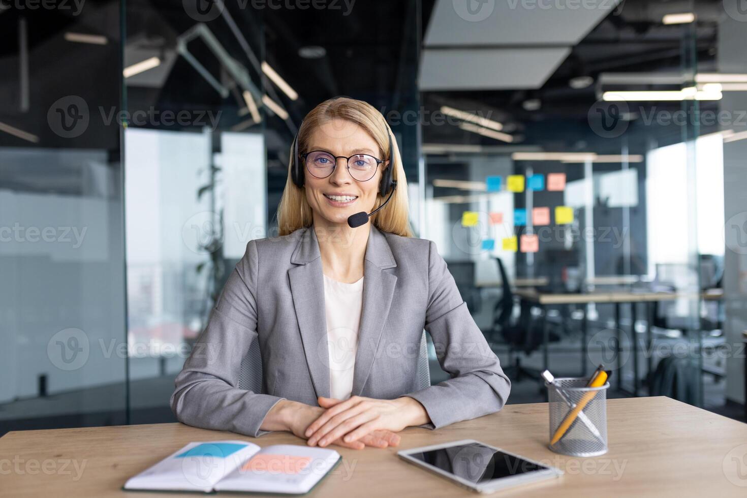A smiling woman in a headset sits at a desk in a modern office environment, ready to assist. She is wearing glasses and a professional attire. photo