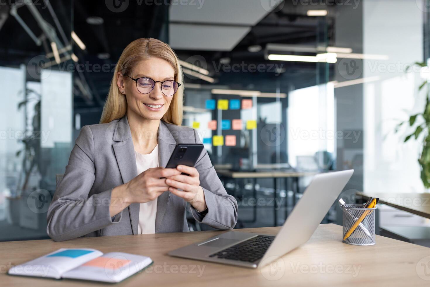 A businesswoman in glasses, dressed in a grey suit, smiles while looking at her phone in a modern office setting with a laptop on a wooden table. photo