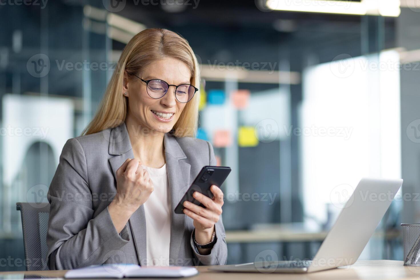 An attractive blonde woman in eyeglasses with a laptop is euphoric about great news she saw on her smartphone in the office. photo