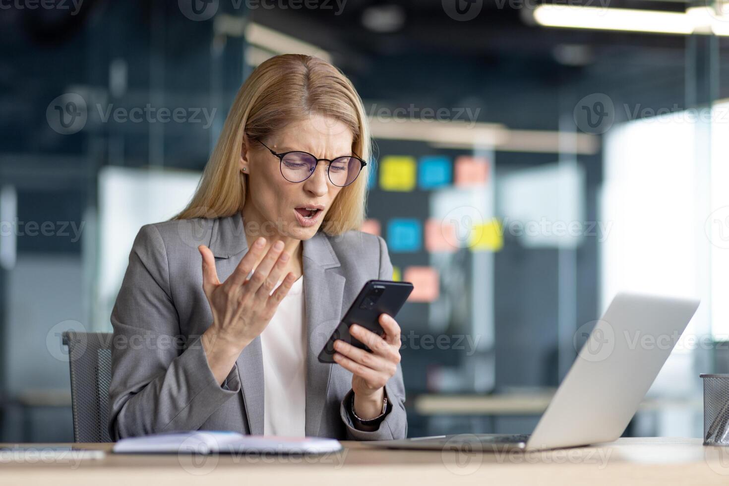 A frustrated businesswoman reacts in surprise while looking at her mobile phone in an office setting. She appears shocked. photo