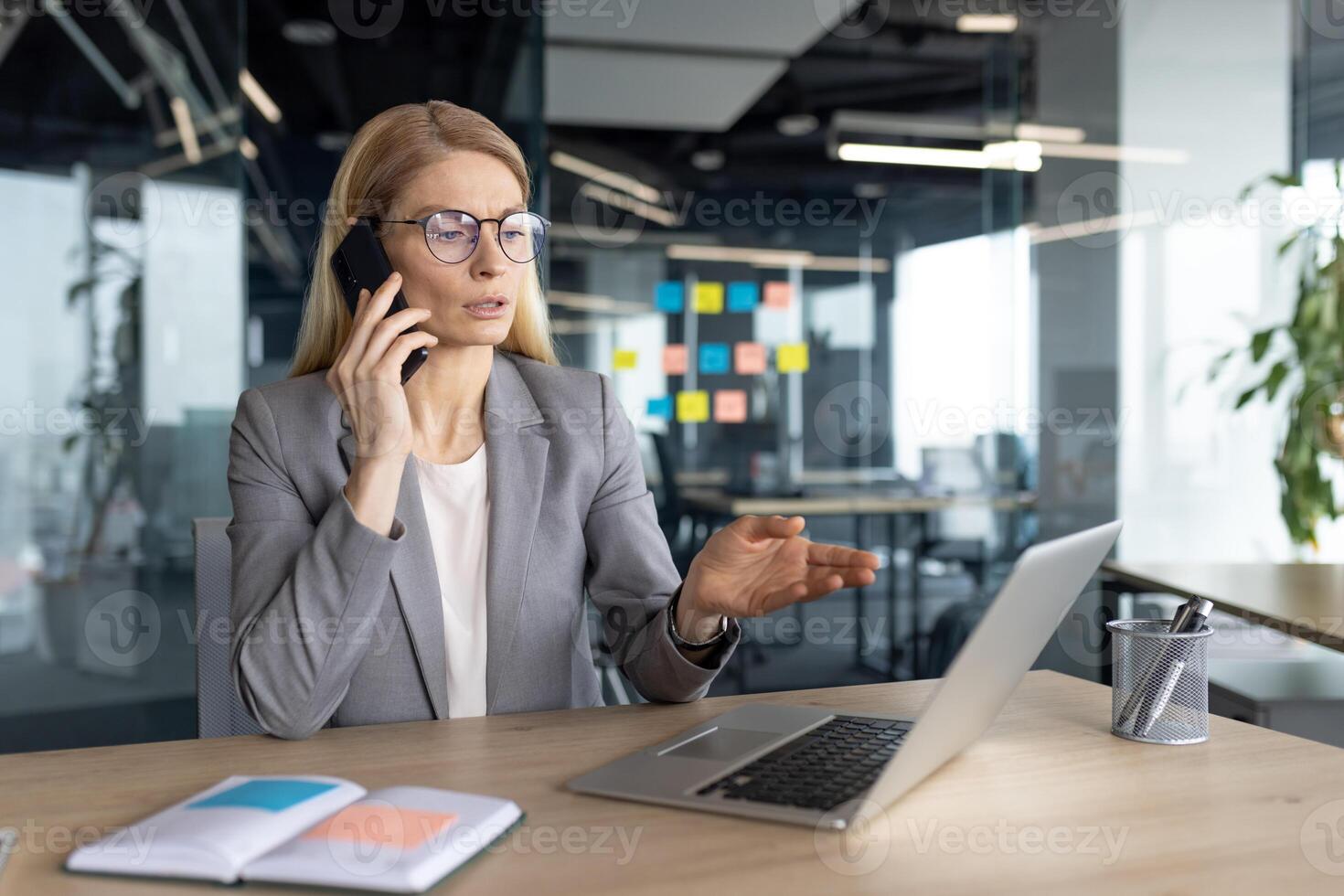 Businesswoman in a grey suit is talking on the phone while gesturing with her hand as she works, at her desk with a laptop in an office. photo