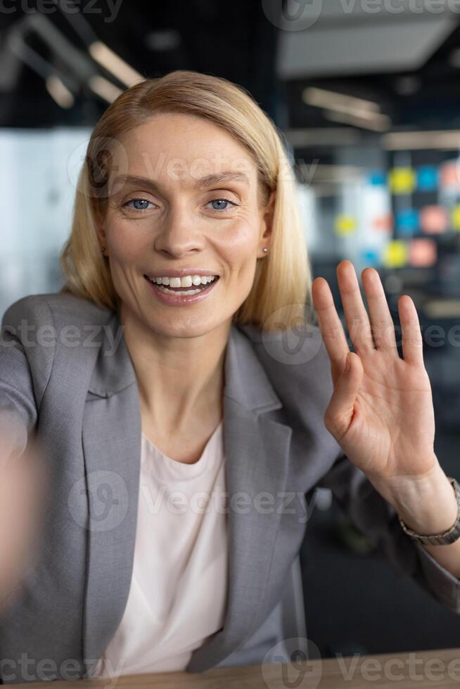 Smiling woman in a grey suit waves hello during a conference call from her office. Focus on positivity and professional connection. photo
