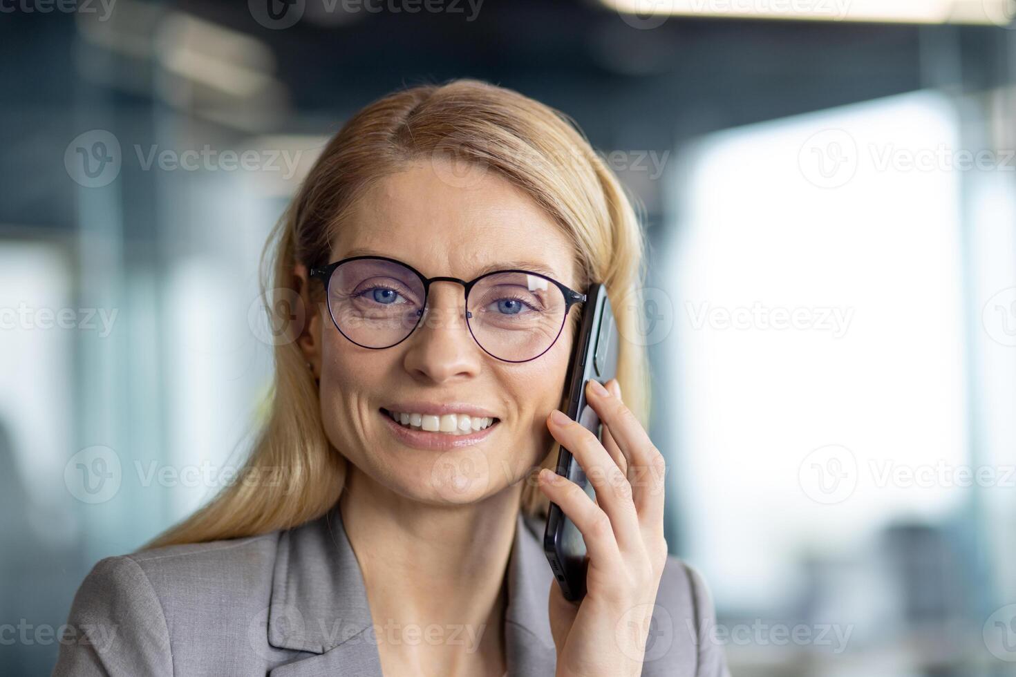 A smiling businesswoman with glasses holds a phone near her ear, looking directly at the camera in a professional setting. photo