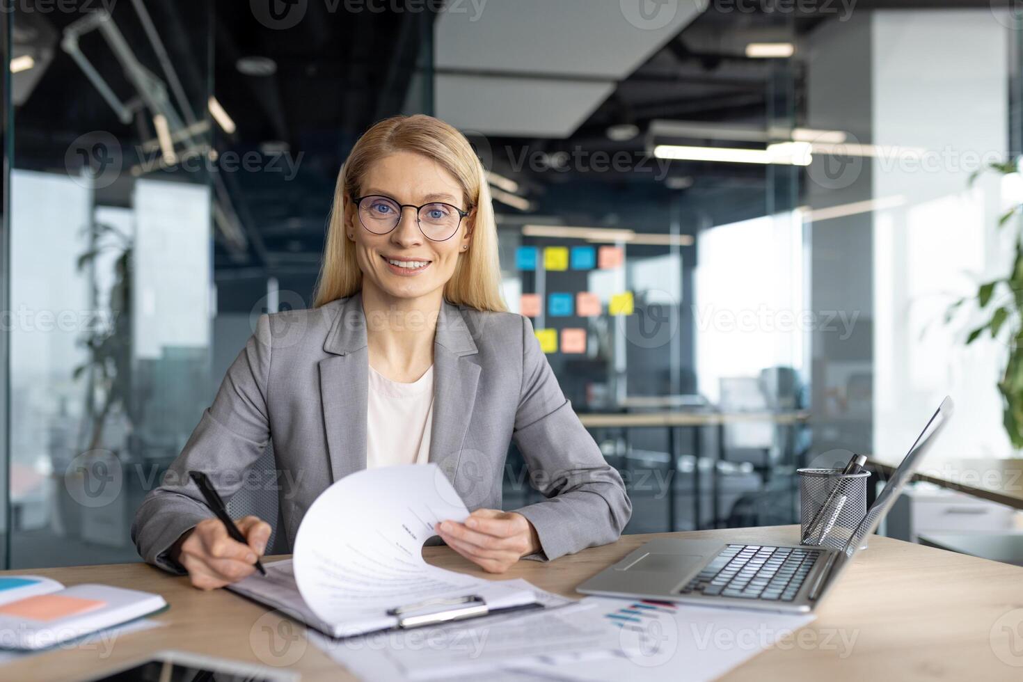A smiling businesswoman, wearing glasses, sits at her desk in a modern office. She is reviewing documents, ready to sign. The laptop is open. photo