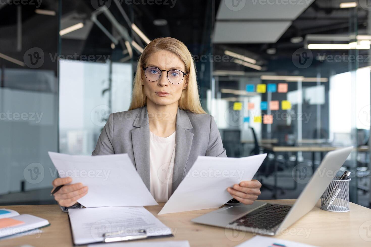 A businesswoman in a professional setting sits at her desk, reviewing documents with a serious expression. She is wearing glasses. photo