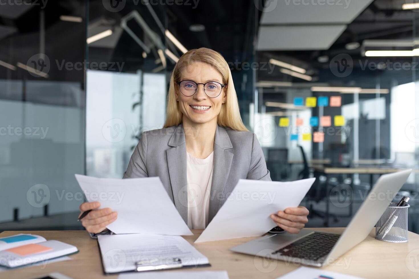 A businesswoman is analyzing documents at her desk with a laptop and smiling in a modern office setting, showcasing her focus and expertise. photo