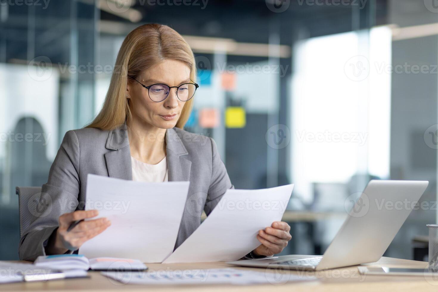A professional woman reviews documents at her desk, using a laptop in a modern office setting. Focus on the business and work. photo