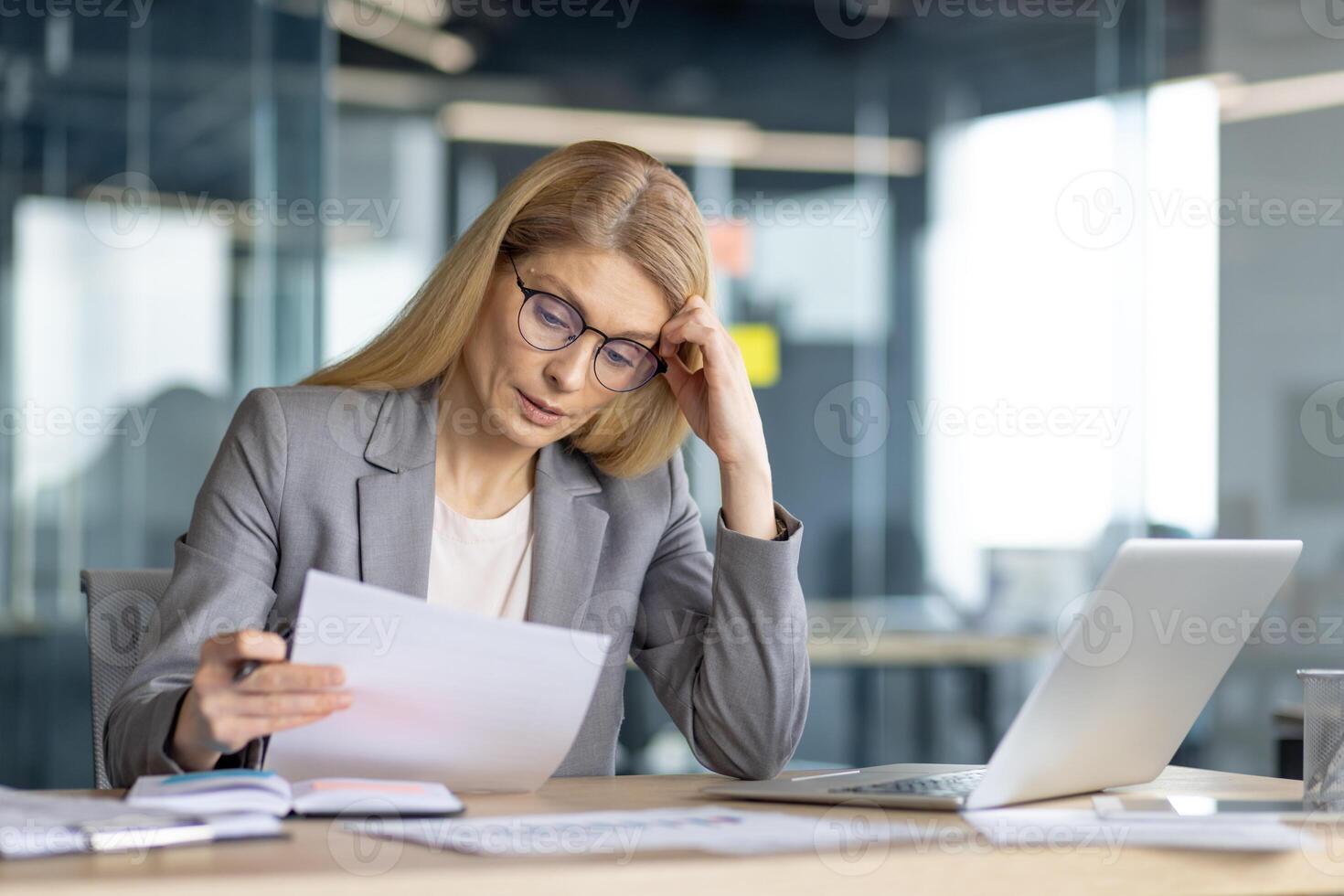 A focused businesswoman, engrossed in the details of paperwork, working in a modern, open office setting with a laptop. photo
