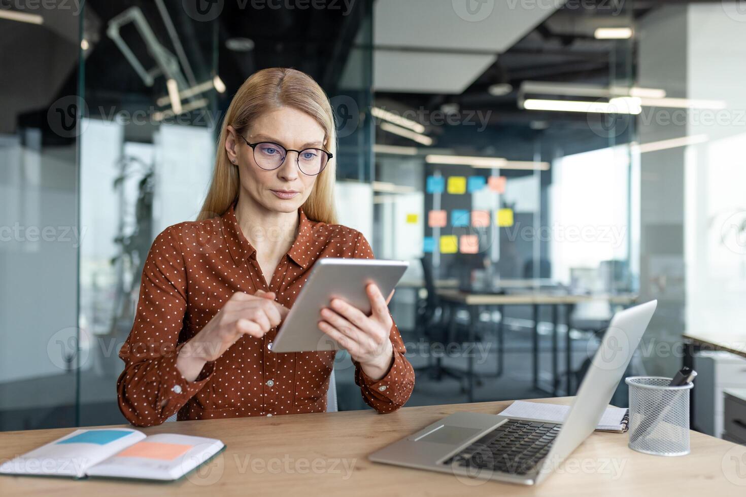 Woman in glasses uses a tablet at her desk, with a laptop and notebook nearby in a modern office setting. photo