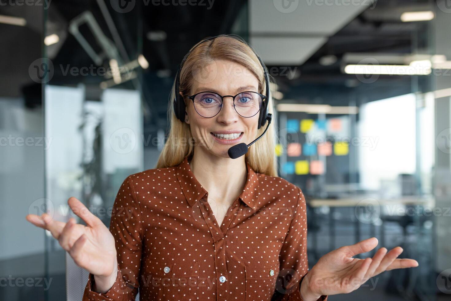 A friendly woman with glasses and a headset smiles while in an office, ready to assist. She gestures with her hands, showing openness and approachability. photo