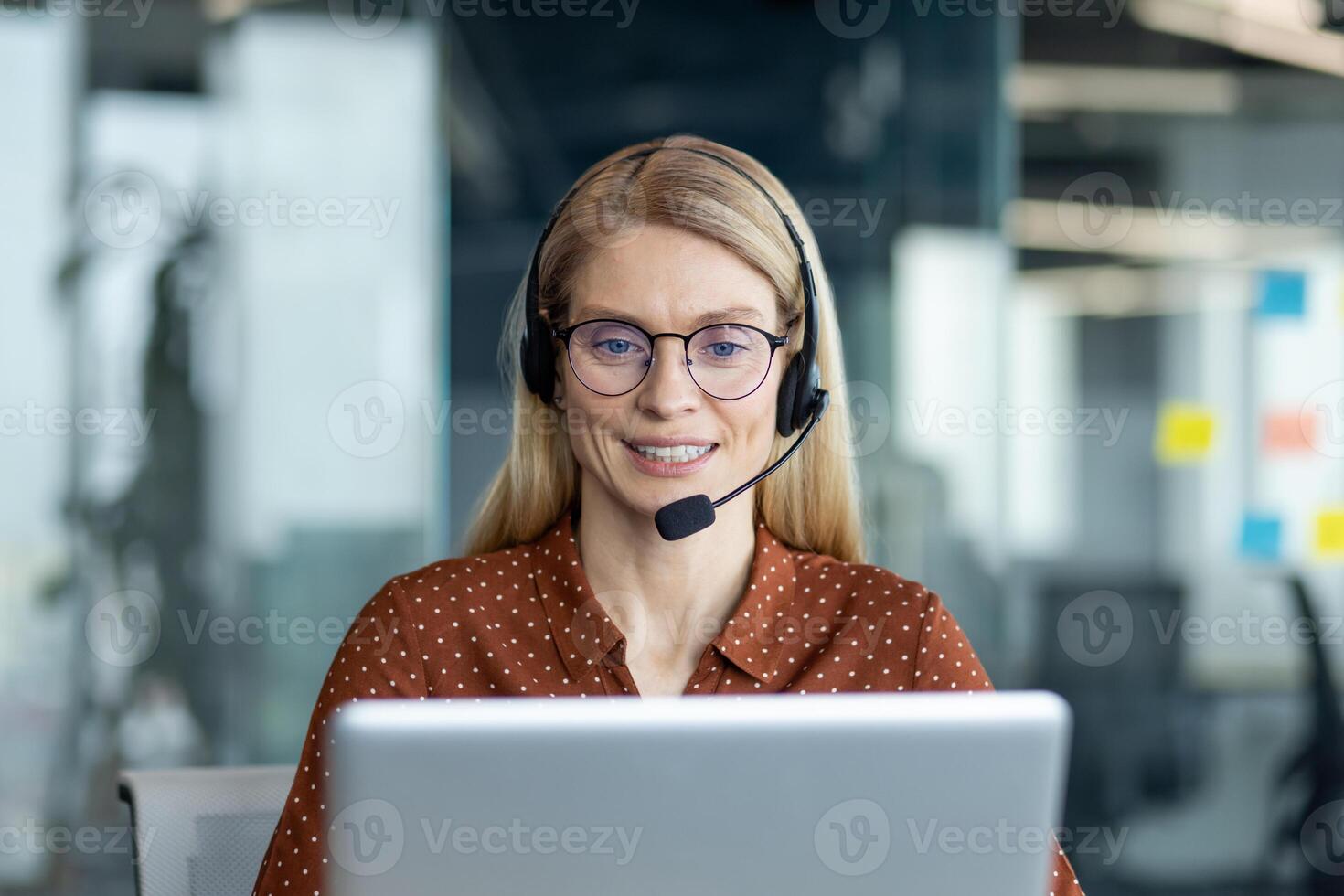 A friendly customer service representative smiles while wearing a headset and working on a laptop at a modern office. photo