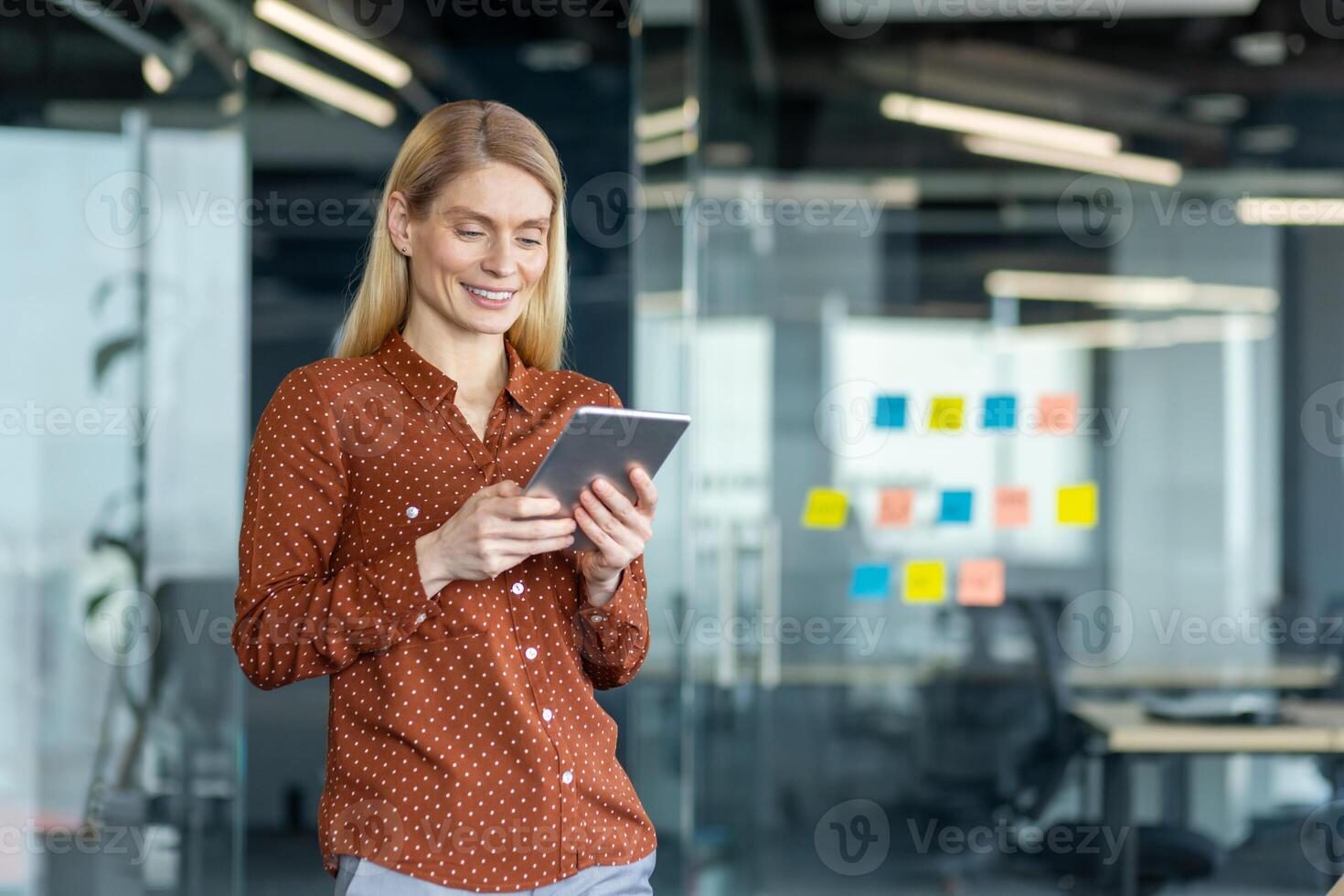 A blonde businesswoman uses a tablet inside a modern office, with glass walls and colorful sticky notes adding a vibrant touch. photo