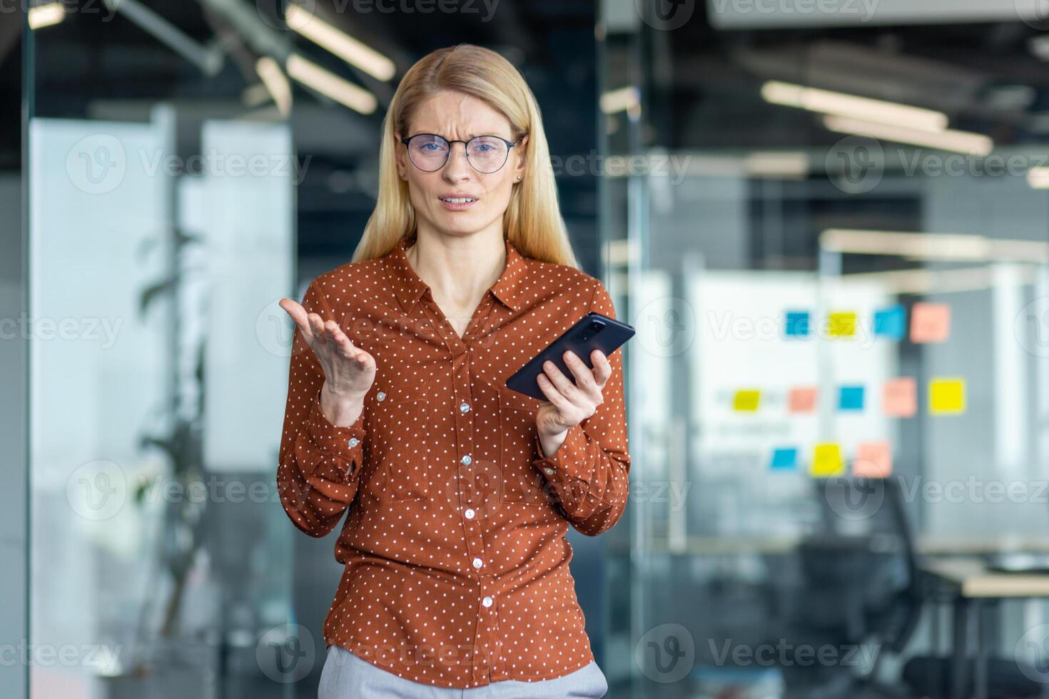 A frustrated woman in glasses holds a phone, gesturing with confusion in an office setting with glass walls and colorful sticky notes. photo