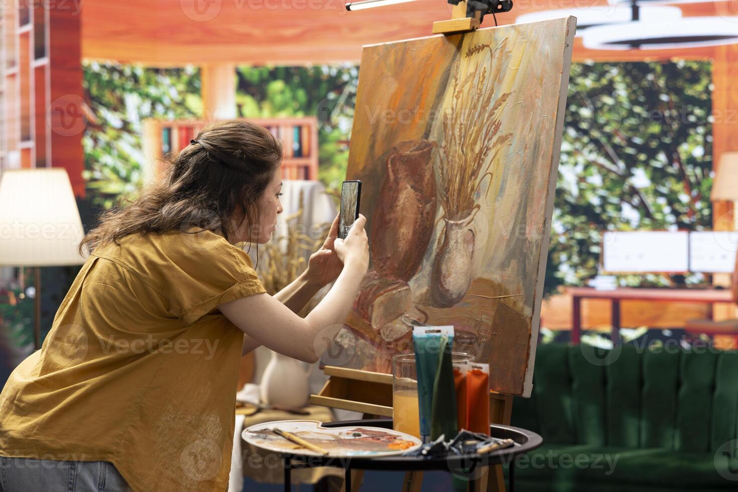 In atelier, painter is promoting her rustic masterpiece painting with the smartphone, creating self advertising content on social media. Filming colorful canvas with fine art piece. Online presence. photo