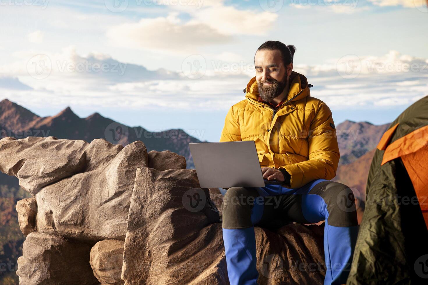 Mountain adventurer prioritizes his online business over safety, working on his laptop despite the extreme winds. The breathtaking landscape is overshadowed by his internet dependency. photo