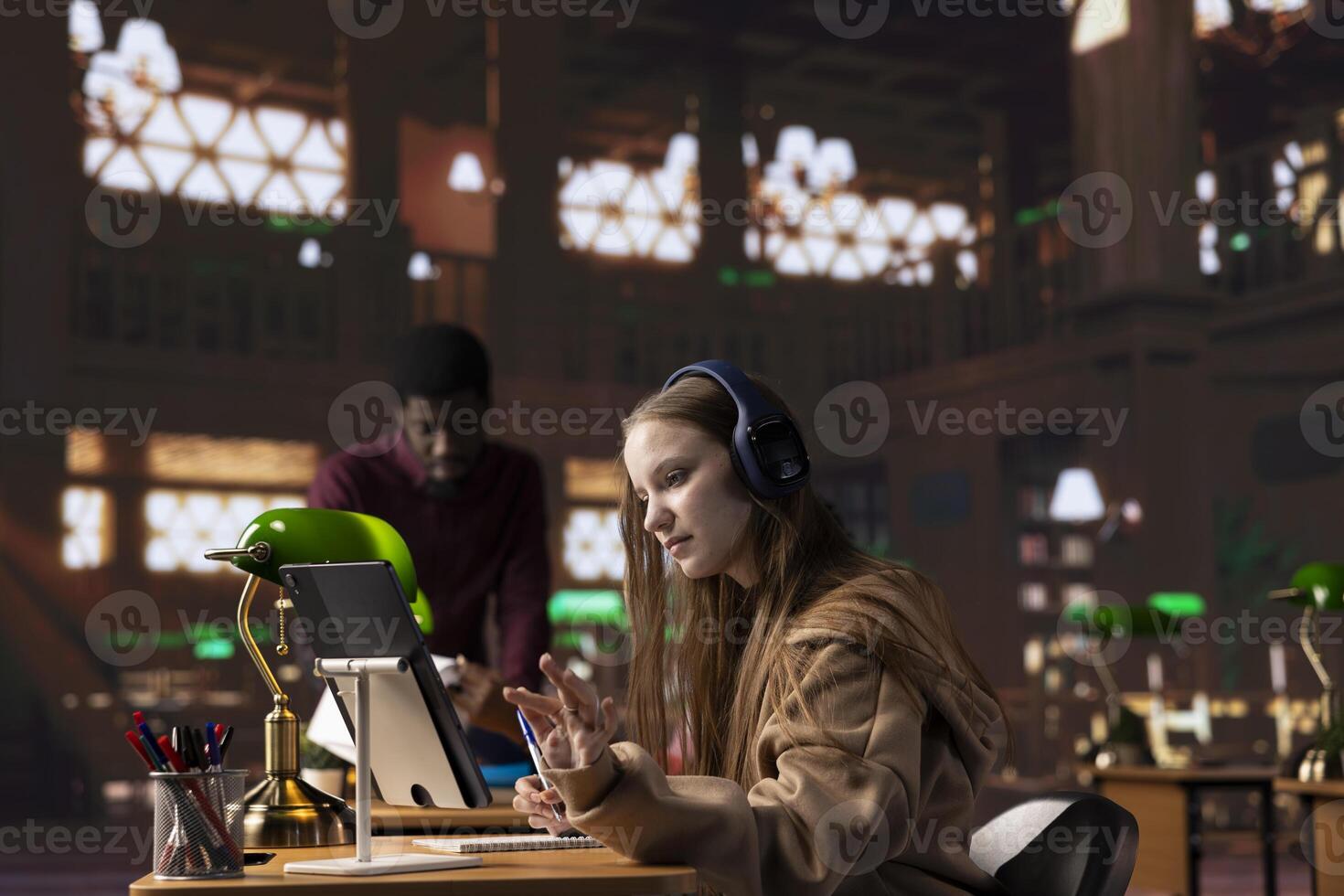 Young girl listening to an educational documentary on her laptop and taking notes, using modern non conventional way to study in the quietness of university library. Student collects academic data. photo