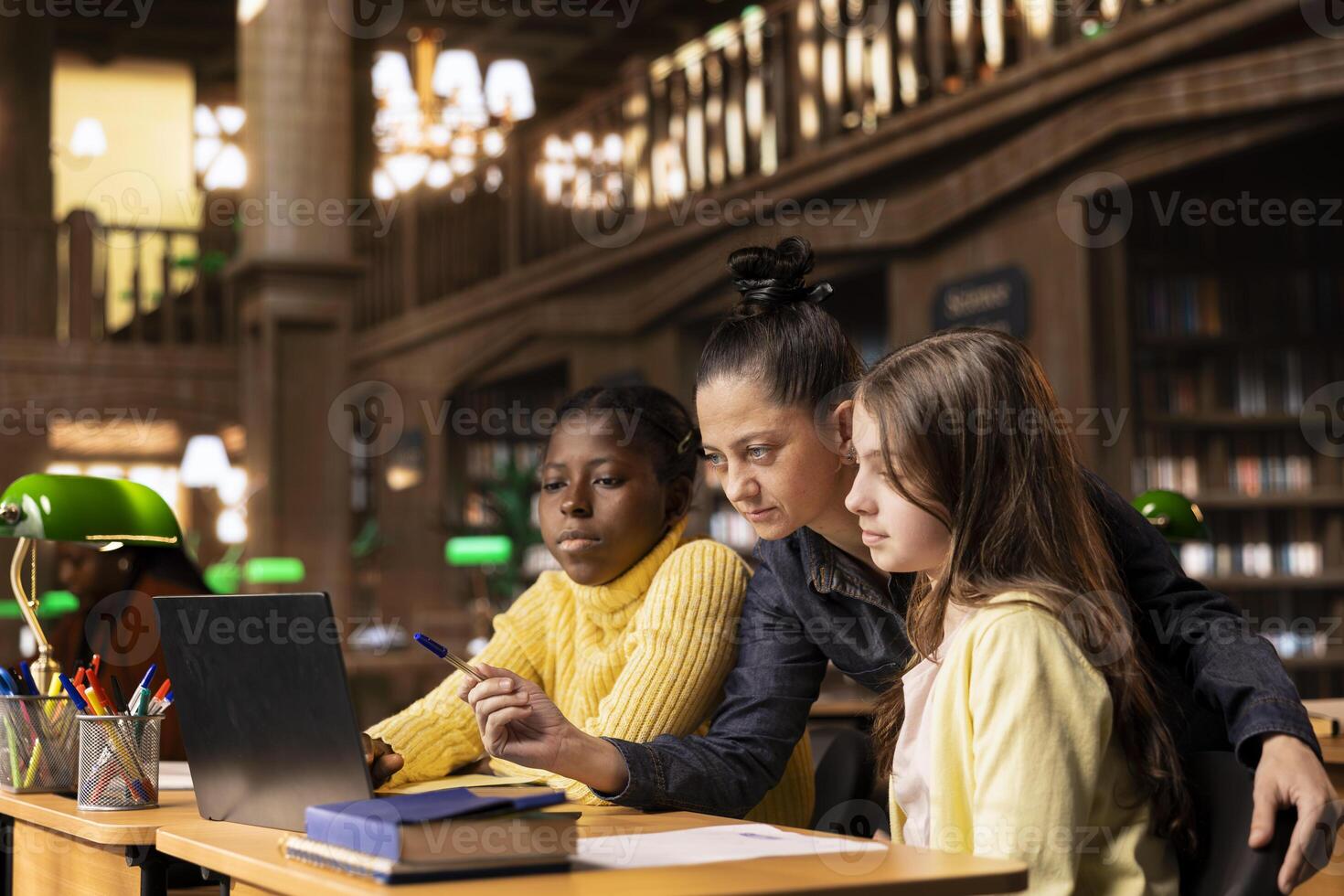 Caucasian tutor guiding two elementary school girls at a library desk, providing private lessons to aid in their academic development with work books and notes. Teacher discussing with scholars. photo