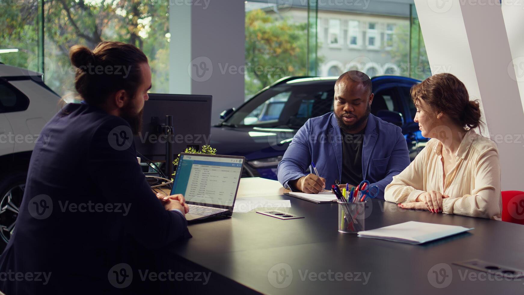 Car dealership salesman sitting with clients at table, giving them contract to sign after finishing test drive. Agent suggesting couple extra vehicle features to buy, waiting for signature, camera B photo